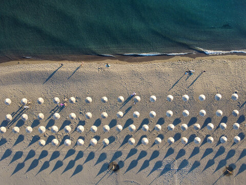 Aerial drone view of a sandy beach resort coastline featuring rows of beach umbrellas casting long shadows on the sand next to the sea. Tourists walking by the sea, view from above.