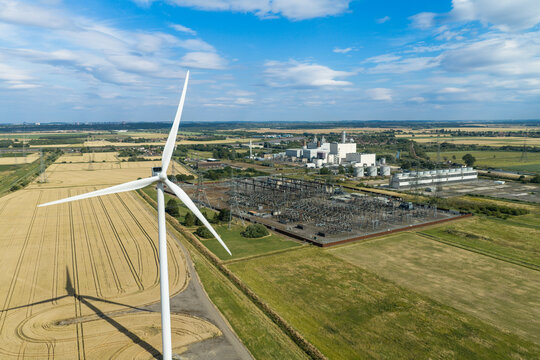 Aerial view of a large wind turbine standing in a golden wheat field, with an industrial power station and electrical substation in the background under a blue sky in the United Kingdom.