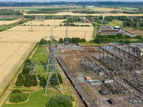 Aerial view of an electrical substation with high-voltage pylons situated amidst expansive agricultural fields and rural landscape in the United Kingdom.