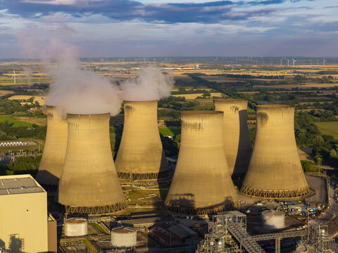 Aerial view of a nuclear power plant with large cooling towers emitting steam, surrounded by green fields and industrial infrastructure in the United Kingdom.