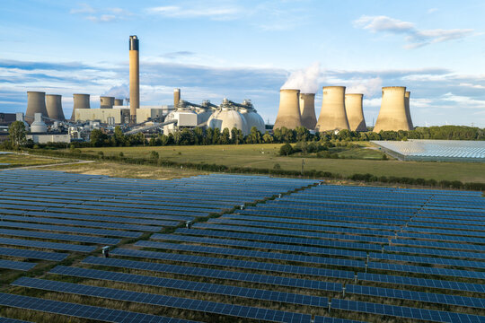 Aerial view of a large solar farm in the foreground with a coal-fired power station and cooling towers in the background under a blue sky in the United Kingdom.