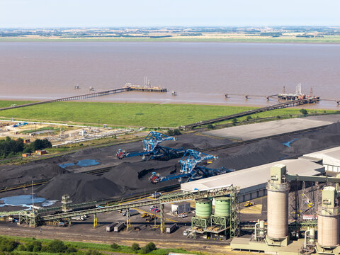 Aerial view of an industrial coal terminal with large stockpiles, heavy machinery, storage silos, and a jetty extending into the river in the United Kingdom.
