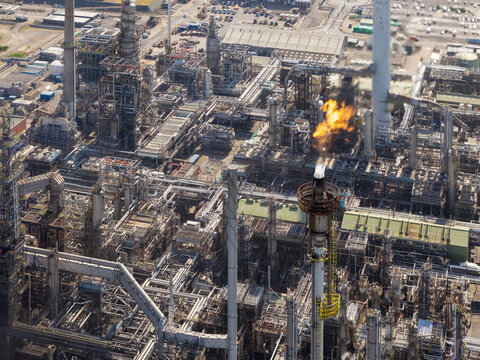 Aerial view of an industrial oil refinery featuring complex piping networks, storage tanks, and a prominent flare stack emitting fire in the United Kingdom.