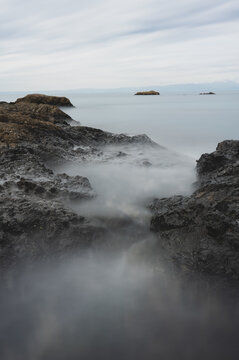 View of dark volcanic rocks along the rugged coastline with misty sea water captured in a long exposure, under a soft overcast sky in Japan.
