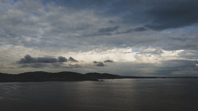 Aerial view of a calm bay bordered by dark rolling hills under a dramatic cloudy sky in Japan.