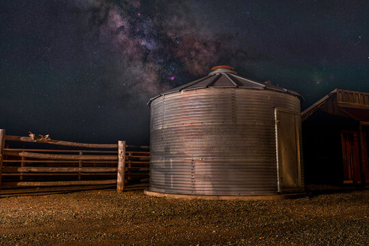 View of a metal harvest silo at a ranch with an old saddle resting on a wooden fence rail under the bright core of the Milky Way galaxy in Picabo, Idaho, United States.