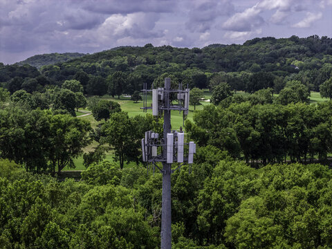 Aerial view of a cell tower equipped with multiple communication antennas, rising above a dense forest canopy and a lush green golf course in Nashville, TN, United States.
