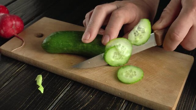 Hand slicing fresh cucumber into thin rounds on a wooden cutting board, with vibrant red radishes and green vegetable scraps visible, creating a colorful kitchen scene