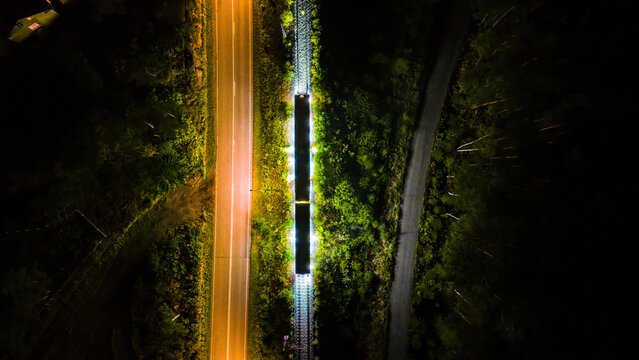 Aerial view of a train illuminated at night traveling on tracks beside a road through a dark forest in Raasepori, Uusimaa, Finland.