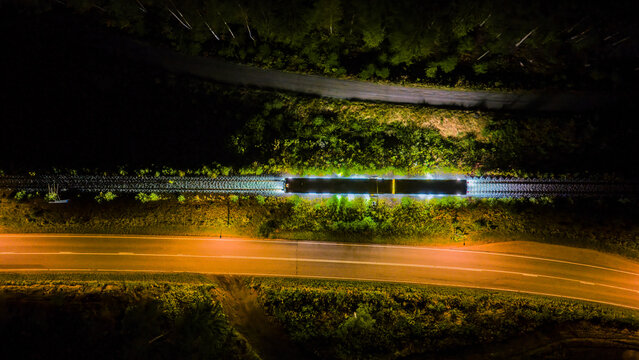 Aerial view of a train illuminated by its own lights traveling along tracks at night, flanked by a dark forest and an adjacent asphalt road in Raasepori, Uusimaa, Finland.