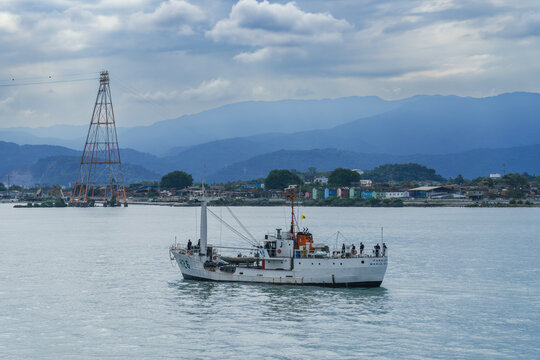 Brazilian Coast Guard ship navigating calm coastal waters with green mountains, power line tower, and overcast sky in the background, daytime maritime patrol scene