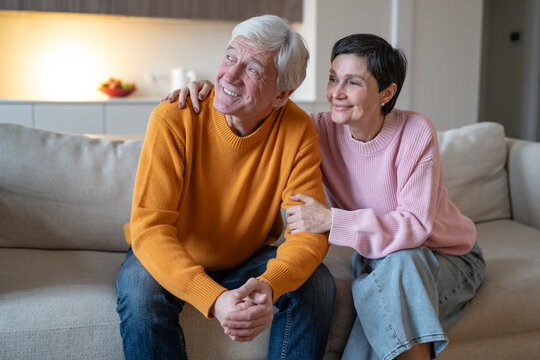 Affectionate cheerful wife hugging smiling husband while couple sitting together on couch at home, joyful positive elderly spouses enjoying conversation and bond, man and woman in happy retirement.