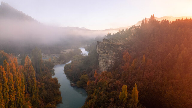 Aerial view of a winding river flowing through a valley with autumn foliage, a prominent rock cliff, and morning mist in Coyhaique, Aysen, Chile.
