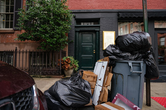 Black garbage bags and cardboard boxes stacked beside a trash bin on a New York City sidewalk. Urban street scene shows residential building entrance and everyday city life.