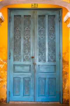 Vintage entrance at building with old closed double wooden doors painted in blue color on yellow wall, ancient wooden door panels with decorative handmade carved patterns and number 24 hanging on top.