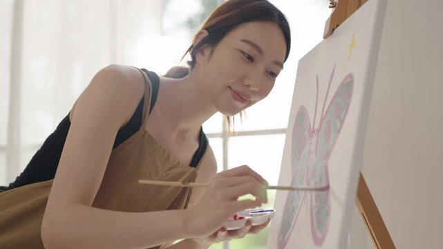 Creative young Asian woman artist painting on canvas in bright home studio. Relaxed female hobbyist enjoying peaceful indoor leisure time, expressing imagination and passion for art.