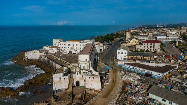 Aerial view of Cape Coast Castle, a historic fortification situated on the rocky coastline of the Gulf of Guinea, surrounded by urban buildings in Cape Coast, Central Region, Ghana.