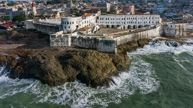 Aerial view of Cape Coast Castle situated on a rocky promontory overlooking the Atlantic Ocean with waves crashing against the shore in Cape Coast, Central Region, Ghana.
