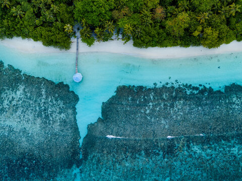 Aerial view of Kihaadhoo island with a wooden pier extending into the turquoise lagoon, surrounded by coral reefs and lush palm trees in Kihaadhoo, Baa Atoll, Maldives.