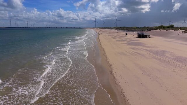 Aerial view video clip of wide sandy beach, North Sea surf waves, coastal sand dunes, wind turbines, and Oosterscheldekering surge barrier in Zeeland, Netherlands on a sunny day