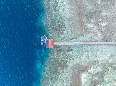 Aerial view of a wooden pier extending over a vibrant coral reef into clear turquoise water with two small boats docked at the end in Malang, East Java, Indonesia.