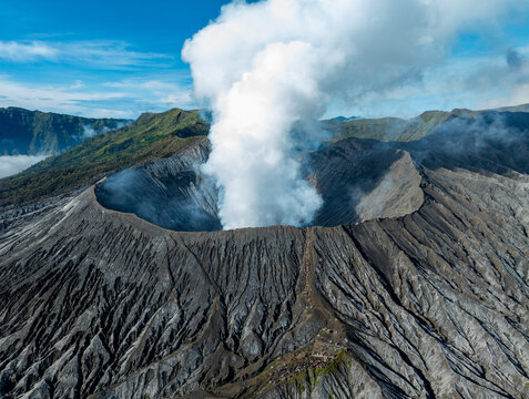 Aerial view of Mount Bromo with its active volcanic crater emitting thick white smoke, surrounded by rugged dark volcanic slopes and distant green hills in Tosari, East Java, Indonesia.