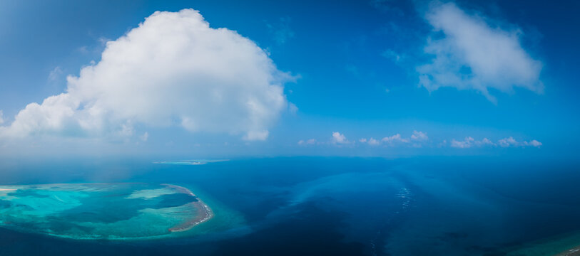 Aerial view of a tropical island surrounded by turquoise coral reefs and deep blue ocean waters under a bright sky with a large white cloud in Dhidhdhoo, Thiladhunmathee Uthur, Maldives.