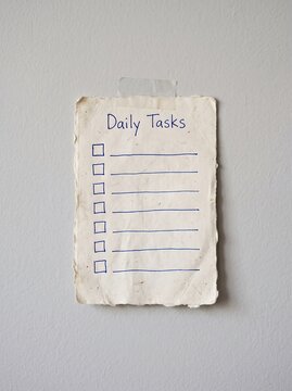 A handwritten daily tasks note on a piece of textured paper taped to a light grey wall