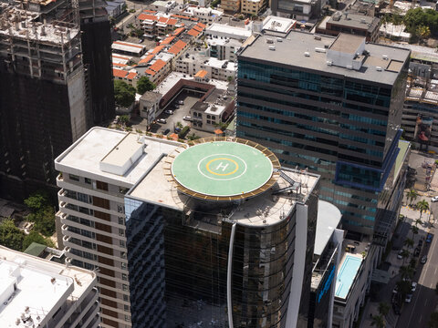 Aerial view of a modern blue glass corporate skyscraper featuring a prominent rooftop helipad structure surrounded by urban buildings and city streets in Santo Domingo, Dominican Republic.
