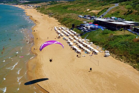 Aerial view of Kisirkaya beach with a paraglider flying over the golden sand, rows of white beach umbrellas, a coastal restaurant, and the blue sea in Sariyer, Istanbul, Turkey.