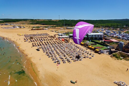 Aerial view of Gumusdere beach with a paraglider soaring above rows of parasols on the sandy shore, with the Black Sea coastline and green hills in Sariyer, Istanbul, Turkey.