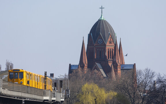 View of a yellow U-Bahn train traveling on an elevated track with the historic St. Thomas Church in the background in Berlin, Berlin, Germany.