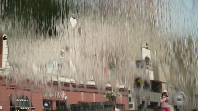 A dense, continuous stream of water flows from the edge of a fountain, shot from inside, with an old architectural building in the background, in the city center, on a hot summer day.