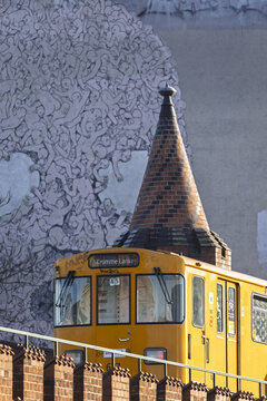 View of a yellow U-Bahn train passing by a historic brick tower and a large mural featuring numerous figures in Berlin, Berlin, Germany.