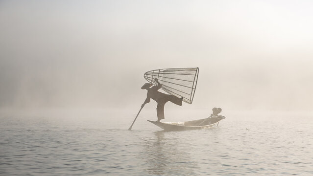 View of a traditional Inle Lake fisherman balancing on a wooden boat while holding a conical net in the misty morning light in Taunggyi, Shan, Myanmar.