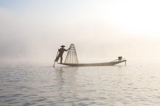 View of an Intha fisherman balancing on a wooden boat with a traditional conical net on the misty waters of Inle Lake in Taunggyi, Shan, Myanmar.