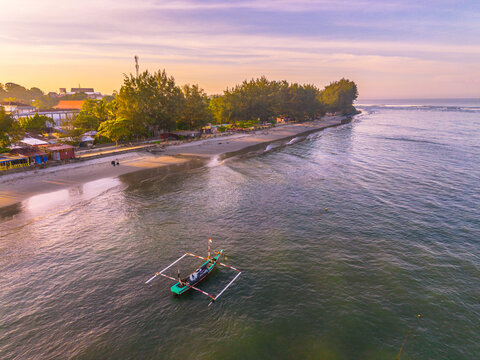 Aerial view of Pantai Panjang Bengkulu with a traditional outrigger boat on the calm sea at sunset, featuring sandy coastline and lush trees in Kecamatan Ratu Agung, Bengkulu, Indonesia.