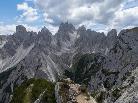 Aerial view of the jagged limestone peaks of the Dolomites with rocky slopes, patches of green vegetation, and dramatic clouds in the sky in Italy.