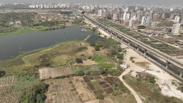 Aerial view of the metro rail line crossing over agricultural fields and a lake near the urban skyline of Dhaka, Dhaka Division, Bangladesh.