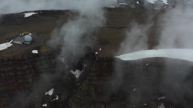 Aerial view of Bolafjall Observation Deck and radar station on the edge of rugged cliffs with patches of snow and low clouds in Bolungarvik, Bolungarvikurkaupstadur, Iceland.