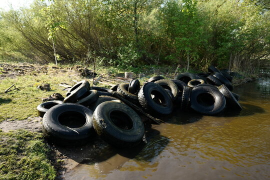 View of a pile of discarded rubber tires on the muddy bank of Rekyva Lake surrounded by lush green trees in Siauliai, Siauliai County, Lithuania.