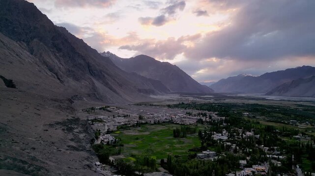 Aerial view of Diskit Monastery nestled in the rugged mountains of the Nubra Valley with lush green fields and dramatic sunset light in Diskit, Ladakh, India.