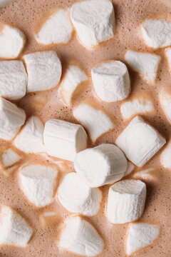 Top view close-up of creamy hot chocolate topped with white mini marshmallows in a white ceramic cup. Cozy sweet winter beverage served on a warm wooden table.