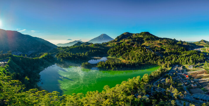 Aerial view of Telaga Warna lake with its vibrant green water, surrounded by lush forested hills, volcanic mountains in the background, and clear blue sky in Kejajar, Central Java, Indonesia.
