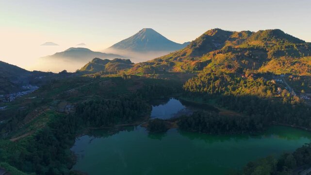 Aerial view of Telaga Warna lake surrounded by volcanic mountains and lush vegetation during golden hour in Kejajar, Central Java, Indonesia.
