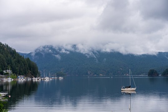 View of a sailboat floating on the calm waters of a bay with a marina and forested mountains under a cloudy sky in Vancouver, British Columbia, Canada.