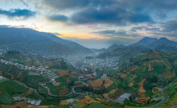 Aerial view of the Dieng Plateau near Telaga Warna, featuring lush terraced fields, winding roads, and a town nestled in the valley in Kejajar, Central Java, Indonesia.