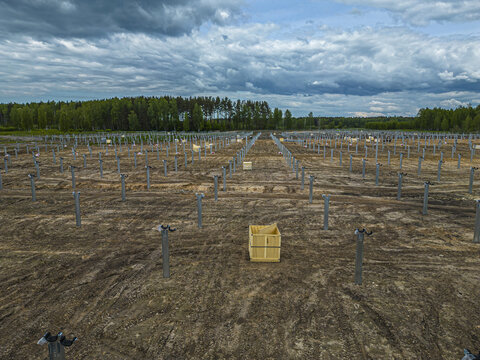 Aerial view of a solar farm construction site with rows of metal support posts installed in the dirt ground, a wooden crate in the foreground, and a forest under a cloudy sky in Riga, Latvia.