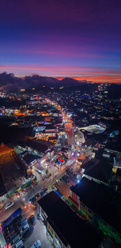 Aerial view of Dieng town at dusk with illuminated streets, traffic light trails, and dense rooftops nestled against a dramatic purple sunset sky. Kejajar, Central Java, Indonesia.