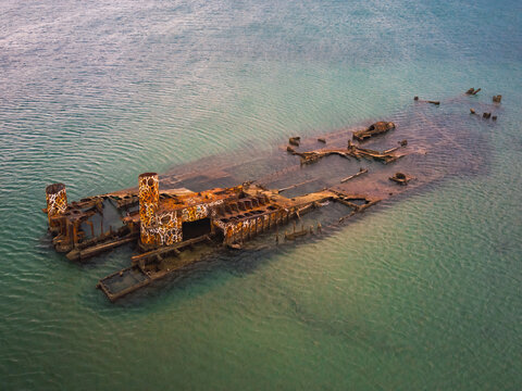 Aerial view of a rusted shipwreck with two vertical towers resting in shallow turquoise water off the coast of Greece.